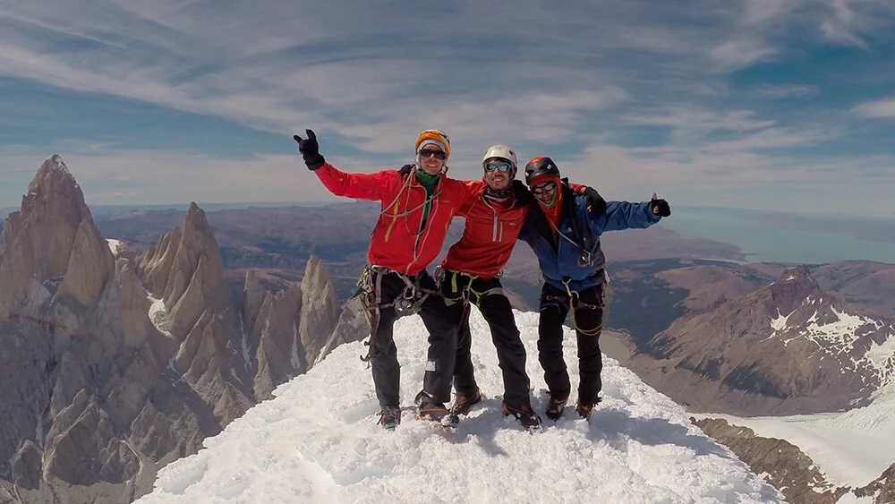 Tres alpinistas en la cima del Cerro Torre — el Fitz Roy y las Agujas al fondo, el Lago Argentino en la distancia