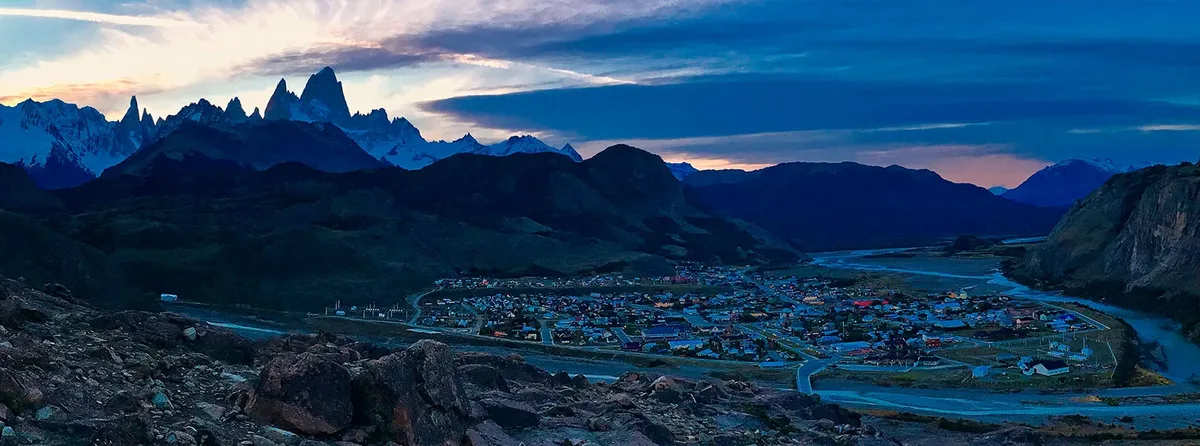 El Chaltén de noche desde las alturas — el pueblo iluminado en el valle con el Fitz Roy y el Cerro Torre recortados en el cielo